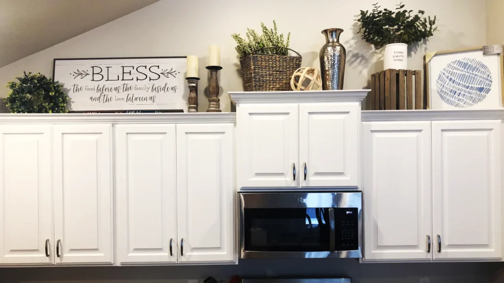 Cluttered mix of small signs, candles, and mismatched decor pieces on top of traditional white kitchen cabinets.